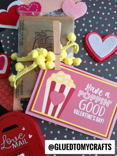 A Valentine’s Day card with a popcorn design and the message “Have a Poppin’ Good Valentine’s Day” is clipped to a bag of popcorn, surrounded by heart decorations and a red mailbox cutout. Perfect as a have a poppin good valentine's day printable!.