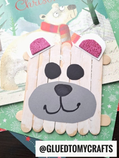 A Christmas craft stick polar bear made from popsicle sticks and paper, with pink glitter ears and a smiling face, is displayed on a festive holiday card background. @GLUEDTOMYCRAFTS is written at the bottom.