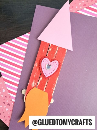 A handmade popsicle stick rocket craft for kids, featuring red painted sticks, a pink paper triangle tip, a pink heart with a gem, orange flames, and layers of pink and purple paper. Text: @GLUETOMYCRAFTS.