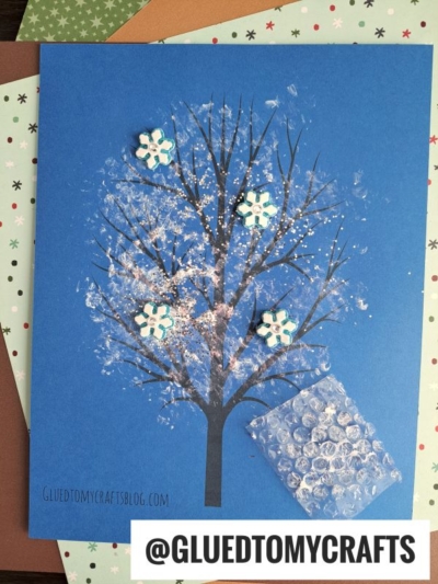 A blue paper shows a painted bare tree with white paint dabbed as snow, creating a bubble wrap snowy tree effect, along with glitter and snowflake decorations. Sheets of scrapbook paper are underneath. The text @GLUETOMYCRAFTS appears at the bottom.