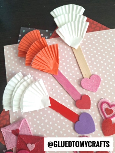 Craft arrows made from popsicle sticks, cupcake liners, and foam heart shapes are displayed on pink heart-patterned paper, capturing a fun cupid's arrow craft perfect for Valentine's Day. Felt hearts and envelopes add a festive touch. Text @GLUEDTOMYCRAFTS is at the bottom.