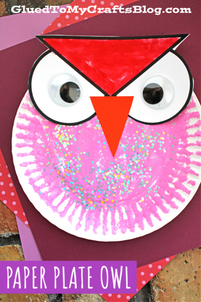 A colorful paper plate valentine owl craft featuring a painted pink plate with glitter, large googly eyes, and a red beak and eyebrows. The background is layered with purple and red paper for extra charm.