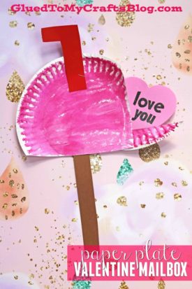A pink and red Paper Plate Mailbox craft with a raised red flag and a heart note that says "love you," displayed on a glittery gold and pink background. Text at the bottom reads paper plate VALENTINE MAILBOX.