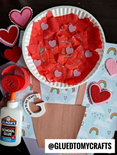 A paper plate hot air balloon is decorated with red tissue paper and glitter, surrounded by heart shapes, glue, and craft materials on a rainbow-themed background. The image features the text @GLUEDTOMYCRAFTS at the bottom.