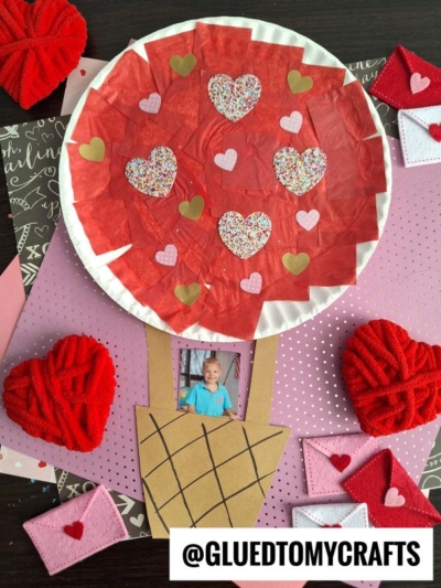 A Valentine’s Day craft shaped like a paper plate valentine hot air balloon, featuring red tissue, heart decorations, and a brown basket with a child’s photo, surrounded by red yarn hearts and envelope cutouts for a festive touch.