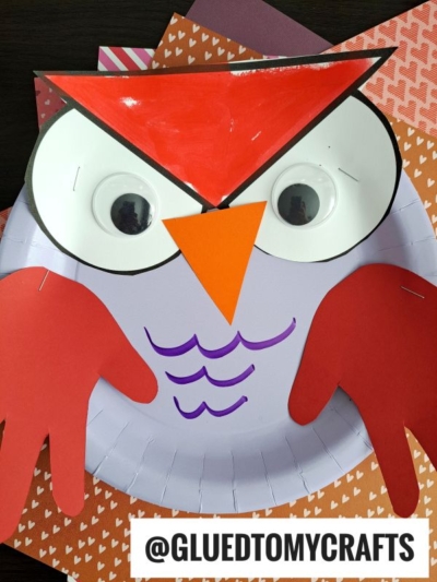A handmade paper plate valentine owl craft featuring large googly eyes, a red and orange paper beak and wings, and purple marker details, placed on heart-patterned paper—a sweet owl kid craft for little ones to enjoy.