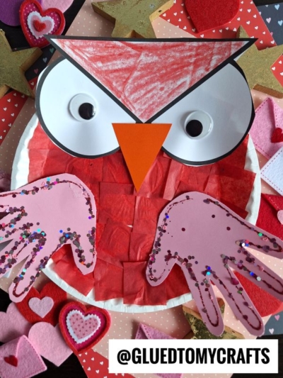 A paper plate valentine owl craft for kids, featuring heart-shaped handprint wings, big eyes, and an orange beak. Surrounded by colorful felt hearts and stars. @GLUEDTOMYCRAFTS is at the bottom.