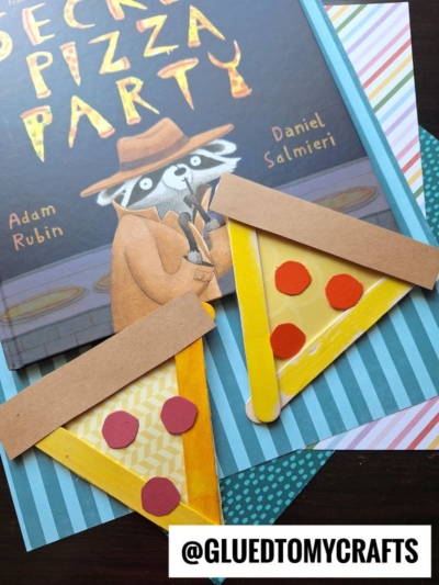 Two popsicle stick pizza slice crafts with colorful paper toppings are placed on top of the book “Secret Pizza Party” by Adam Rubin and Daniel Salmieri, along with striped patterned paper. @gluedtomycrafts is written at the bottom.