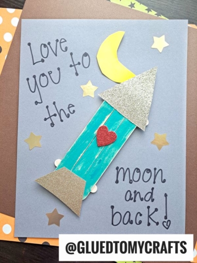 A handmade card with a popsicle stick valentine rocket ship, gold stars, and a crescent moon, reading “Love you to the moon and back!” The rocket has glittery silver details and a red heart. @GLUEDTOMYCRAFTS is written at the bottom.