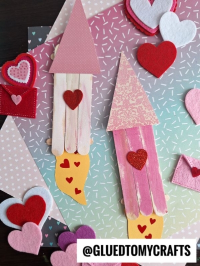 Two popsicle stick valentine rocket ships, made with colorful paper and felt hearts, are displayed on a pastel, patterned background with heart decorations. A fun valentine rocket craft for kids! The Instagram handle @GLUEDTOMYCRAFTS appears at the bottom.