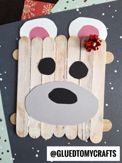 A craft stick polar bear face made from colored paper, with a red bow on one ear, is displayed on a dark background with snowflake patterns. Text reads @GLUEDTOMYCRAFTS at the bottom.