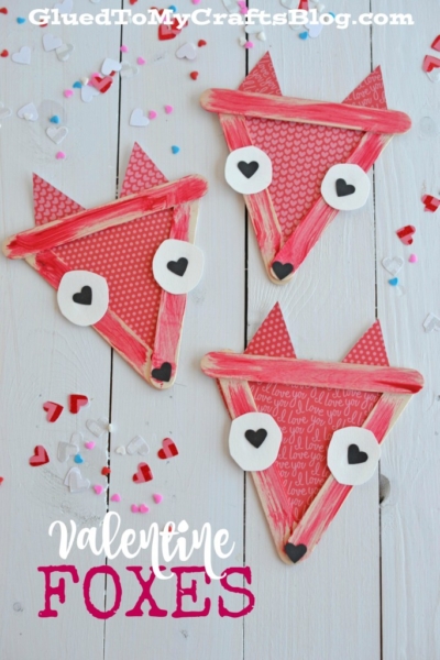 Three red and pink popsicle stick valentine fox faces made from craft sticks, paper, and heart shapes are arranged on a white wooden surface with scattered confetti. Text reads Valentine Foxes at the bottom.