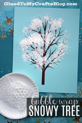 A handmade bubble wrap snowy tree art on blue paper features painted branches with white paint snow dabbed using bubble wrap, displayed on a plate; gold stars shine in the background. Text reads, “bubble wrap SNOWY TREE.”.