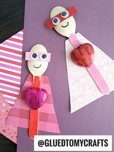 Two wooden spoon valentine superheroes with paper capes, red and pink masks, googly eyes, drawn smiles, and heart-shaped chocolates on their chests are displayed on pink scrapbook paper.