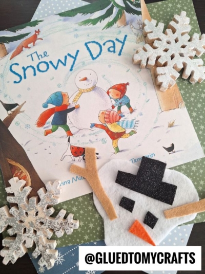 A childrens book titled The Snowy Day with illustrated children and a snowman, two decorative snowflakes, a felt melted snowman face, and the text @GLUEDTOMYCRAFTS are arranged on a table.