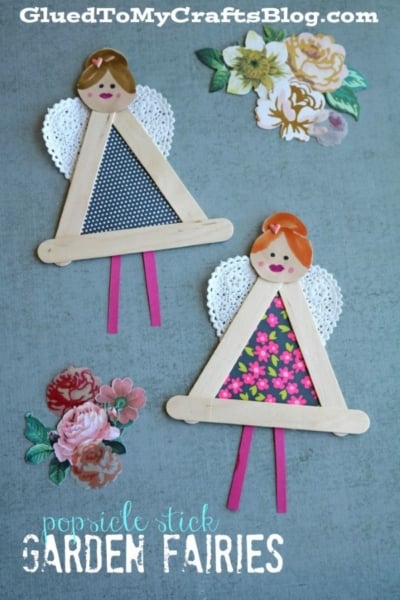 Two handmade popsicle stick garden fairies are crafted from popsicle sticks, patterned paper, and delicate lace wings, each decorated with painted faces. They are set on a gray surface adorned with floral stickers. Text reads: “popsicle stick GARDEN FAIRIES.”.