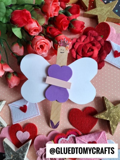 A popsicle stick valentine fairy, crafted from white and purple paper with a hand-drawn face, sits among red roses, felt hearts, stars, and paper envelopes. The handle @GLUEDTOMYCRAFTS is visible at the bottom.