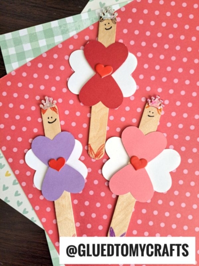 Three popsicle stick valentine fairies with heart-shaped wings and crowns, each sporting a smiley face, are arranged on polka-dot and checked paper backgrounds. The handle @GLUEDTOMYCRAFTS appears at the bottom.