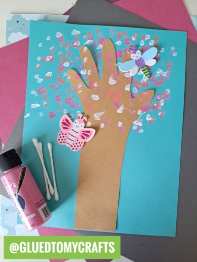 A kids’ craft featuring a Handprint Cherry Blossom Tree made from a brown paper hand cutout on blue paper, decorated with q-tip cherry blossom paint spots, two foam butterfly stickers, and pink paint. @GLUEDTOMYCRAFTS is labeled at the bottom.