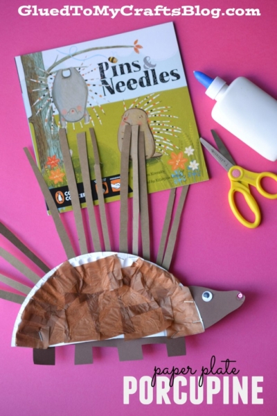 A porcupine paper plate craft made from a paper plate, brown paper, and construction paper sits on a pink surface near scissors, glue, and the book Pins & Needles, with paper plate porcupine text below.