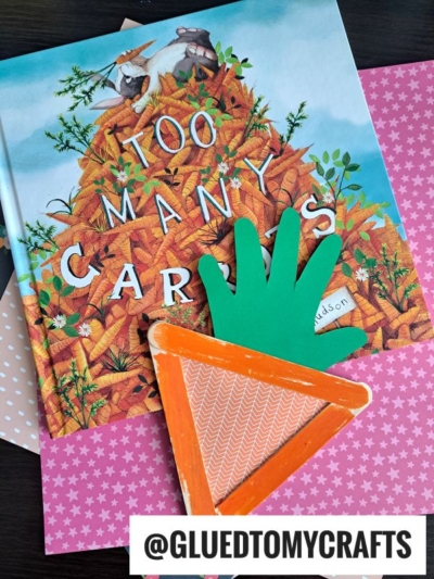 A children’s book titled Too Many Carrots lies on pink and peach patterned paper. On top, there’s a carrot-shaped craft made from an orange triangle with a green paper hand for leaves—perfect for popsicle stick carrots fun! Tag reads @GLUEDTOMYCRAFTS.