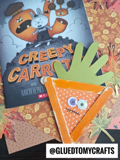 A children’s book titled Creepy Carrots is shown with a handmade carrot craft made from popsicle stick carrots and paper, placed on a surface decorated with paper leaves. The handle @GLUEDTOMYCRAFTS is visible.