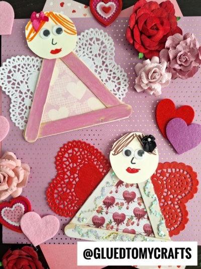 Two craft stick garden fairies with lace doily wings are displayed on a pink polka-dot background, surrounded by paper flowers and felt hearts. The image includes the text @GLUEDTOMYCRAFTS at the bottom.