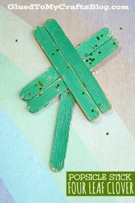 Four green craft sticks arranged in a four-leaf clover shape, decorated with green glitter on a pastel rainbow background. Text reads Popsicle Stick Clover.