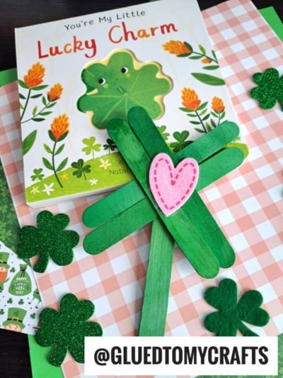 A children’s book titled “You’re My Little Lucky Charm” lies on a checkered surface, surrounded by shamrock decorations and a Popsicle Stick Clover crafted with a pink heart in the center.