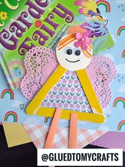 A colorful handmade popsicle stick garden fairy craft made from paper, a doily, popsicle sticks, and decorative items sits on a book titled Garden Fairy, with a pastel rainbow-themed background. @GLUEDTOMYCRAFTS is written at the bottom.