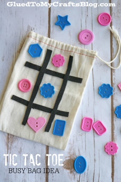 A cloth bag with a black tape tic-tac-toe grid and blue and pink button pieces, perfect for an on-the-go tic tac toe game. More buttons are scattered on a wooden surface. Text reads Tic Tac Toe Busy Bag Idea.