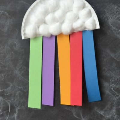A paper plate rainbow craft features cotton ball clouds and colorful paper strips—green, purple, yellow, red, and blue—dangling like a rainbow. This cheerful rainbow cloud craft hangs on a string against a dark background.