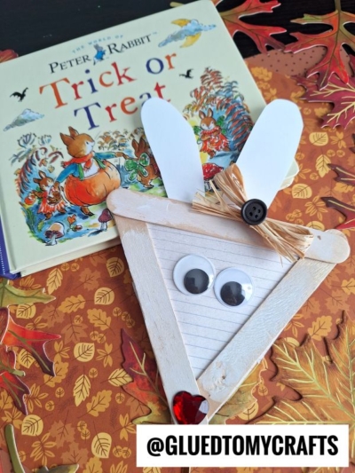 A Peter Rabbit Trick or Treat book and a charming handmade bunny face—crafted from popsicle sticks, googly eyes, a button, and a heart gem—rest on a cozy fall-themed background. This delightful bunny popsicle stick craft adds extra whimsy to the scene. @GLUEDTOMYCRAFTS label at the bottom.