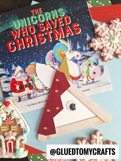 A Christmas craft scene with a book titled The Unicorns Who Saved Christmas, Popsicle Stick Unicorns, and decorated cookies shaped like snowflakes and a gingerbread house. @GLUEDTOMYCRAFTS tag is visible.