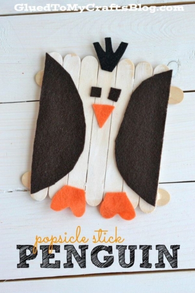 A popsicle stick penguin craft made from black and orange felt for the wings, feet, and beak, arranged on a white wooden surface.