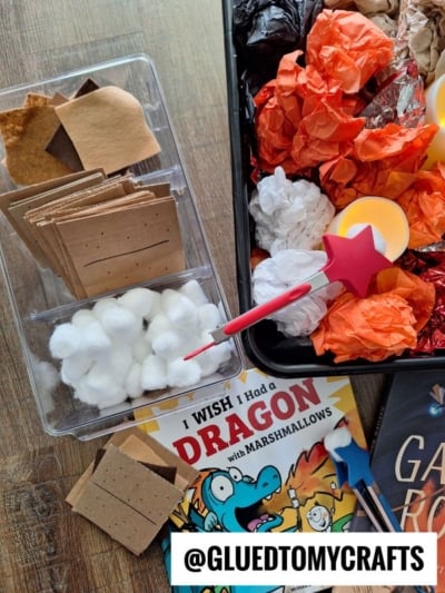A tabletop displays a “Dragon with Marshmallows” craft setup: tissue paper (red, orange, black, white), flameless candles for pretend play s’mores, cotton balls, graham crackers, a dragon book, magic wand, and @gluedtomycrafts label.
