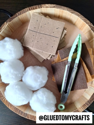 A wooden plate holds pretend play smores, with cotton balls, brown felt squares, cardboard squares with dots, and mint-green tongs. A label at the bottom reads @GLUETOMYCRAFTS.