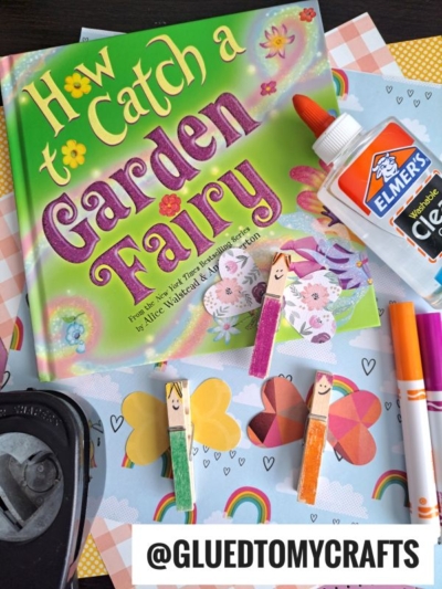 A childrens craft setup featuring the book How to Catch a Garden Fairy, clear Elmers glue, markers, paper, a hole punch, and three adorable clothespin fairies with fairy faces and paper wings. @GLUEDTOMYCRAFTS.