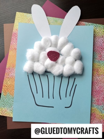 A handmade cotton ball bunny cupcake craft features fluffy cotton balls as frosting, pink glitter paper as a nose, and white paper bunny ears on blue paper, set atop colorful sheets. @GLUEDTOMYCRAFTS is written at the bottom.