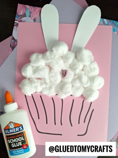 A cotton ball bunny cupcake craft with paper bunny ears and a heart gem sits on colored paper beside Elmer’s school glue. The adorable bunny cupcake creation is shared by @GLUEDTOMYCRAFTS at the bottom.