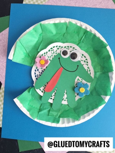 A Paper Plate Lily Pad craft featuring a green frog made from a hand-shaped cutout, googly eyes, flowers, and a red tongue, displayed on blue and green paper. @GluedToMyCrafts is written at the bottom.