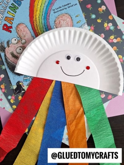 A Paper Plate Rainbow craft shaped like a smiling rainbow, with googly eyes, a drawn mouth, and red, blue, yellow, and green crepe paper strips hanging down—perfect for pairing with a cotton ball rainbow cloud or your favorite rainbow-themed book.