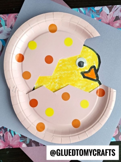 A paper plate hatching baby chick craft featuring a yellow chick with a googly eye peeking from a pink, polka-dotted eggshell. The background has floral patterns and colored paper. @GLUEDTOMYCRAFTS is written at the bottom.