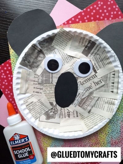A paper plate newspaper koala craft with newspaper pieces, large googly eyes, and black paper ears and mouth. A bottle of Elmers School Glue lies beside it, with a “@GLUEDTOMYCRAFTS” label at the bottom right.
