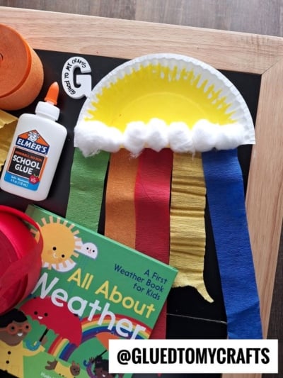 A colorful paper plate rainbow craft features a cotton ball cloud and strips of red, blue, yellow, and green crepe paper. Nearby are a glue bottle, extra crepe paper, and a children’s weather book for inspiration.