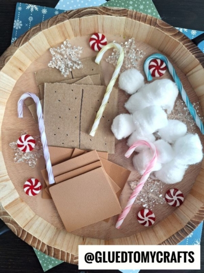 A wooden tray holds candy canes, cotton balls, red-and-white peppermint candies, faux snowflakes, and tan foam rectangles resembling graham crackers for a winter pretend play smores craft. The handle @GLUEDTOMYCRAFTS is visible at the bottom.