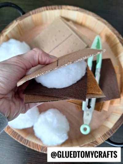 A hand holds a pretend play smore made of brown felt, cardboard, and white cotton, with more crafting materials, cotton balls, and green scissors in a wooden tray. Text at the bottom reads @GLUEDTOMYCRAFTS.