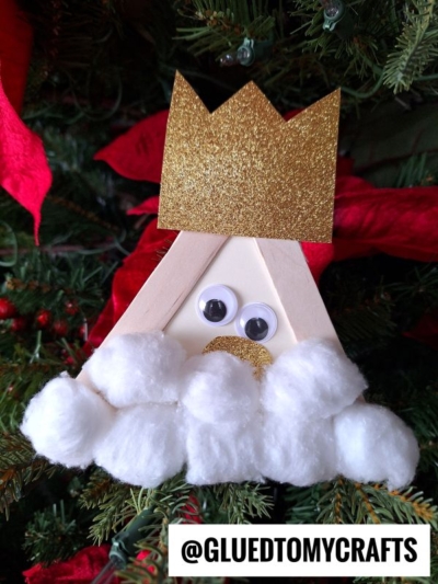 A craft stick king made from popsicle sticks, with a gold glitter crown, googly eyes, and a big cotton ball beard, hangs on a Christmas tree. The username @GLUEDTOMYCRAFTS appears at the bottom. Perfect for popsicle stick kings crafts!.