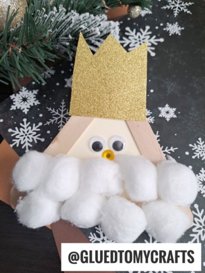 A handmade popsicle stick king craft featuring a gold glitter crown, googly eyes, yellow nose, and a cotton ball beard, set on a black background with white snowflake patterns. @GLUEDTOMYCRAFTS is written at the bottom.