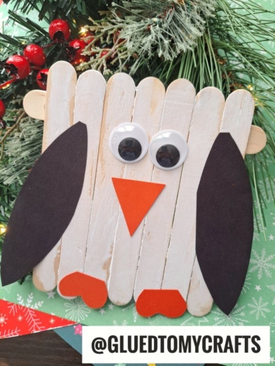 A DIY owl craft made from painted popsicle sticks, black paper wings, orange paper beak and feet, and two large googly eyes—just like a festive craft stick penguin—placed near Christmas greenery and decorations. @GLUEDTOMYCRAFTS text at the bottom.